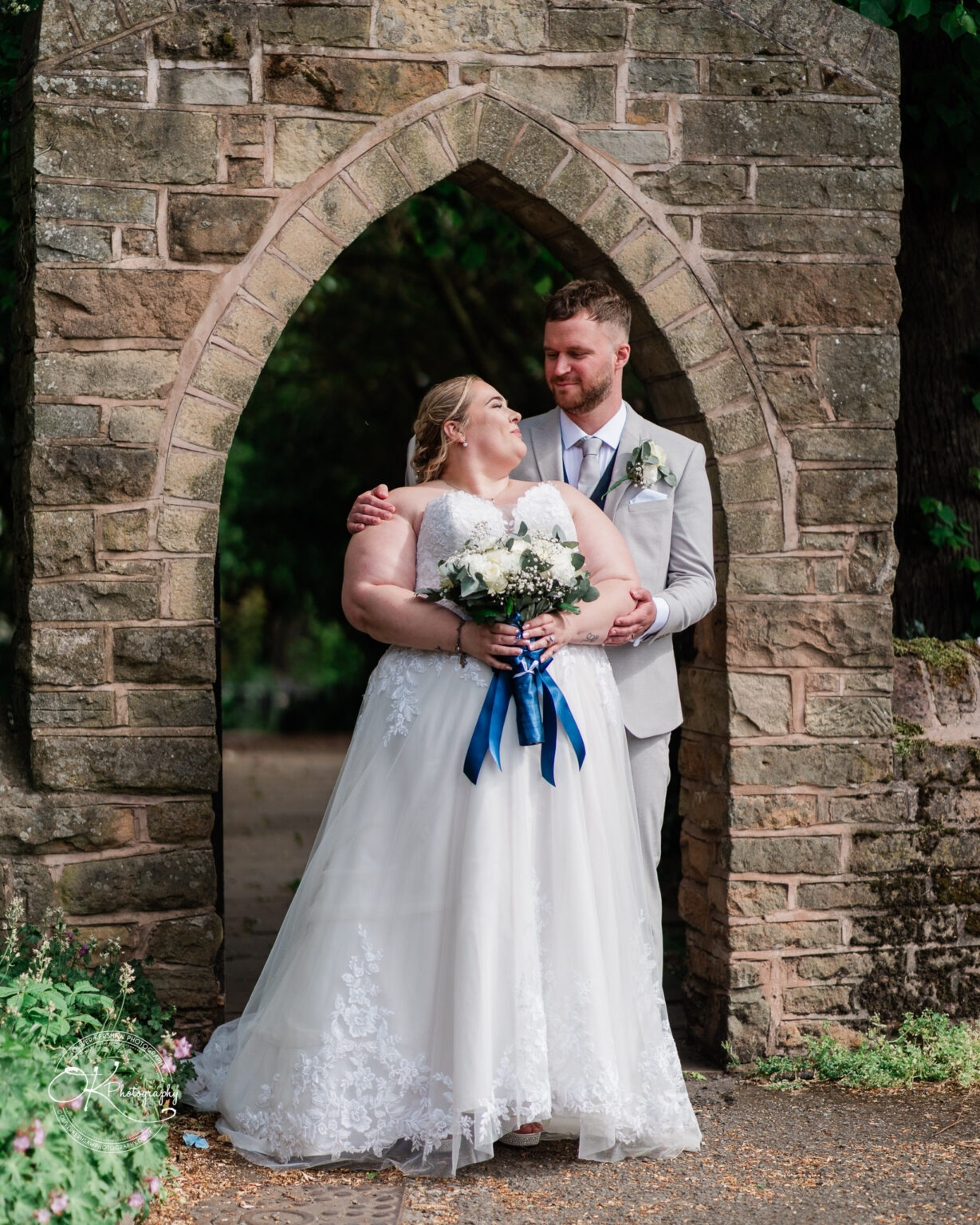 A bride and groom embrace under a stone archway, the bride holding a bouquet with white flowers and blue ribbons.