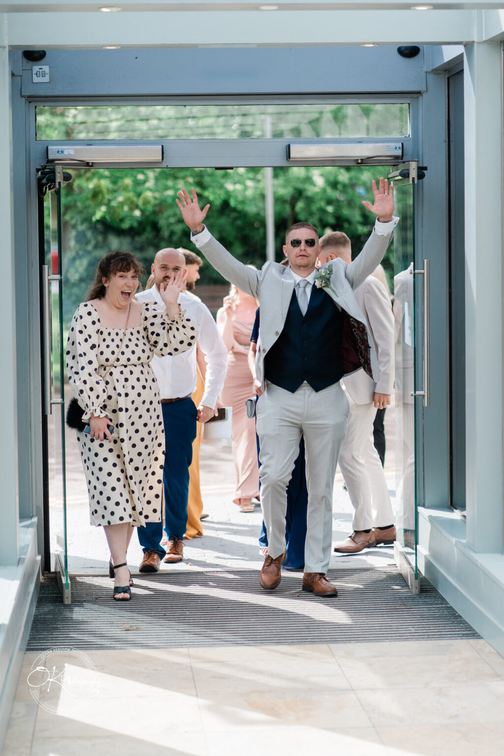 A groom in a light-coloured suit raises his arms joyfully at the doorway, while guests, including a woman in a polka dot dress, wave.
