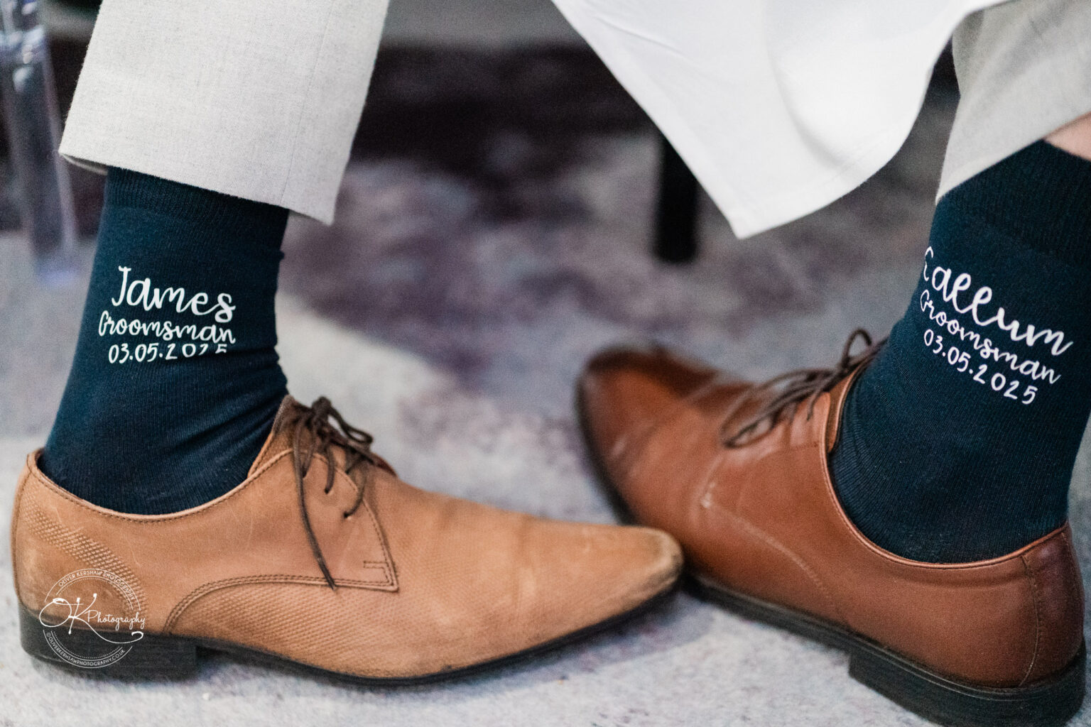 Brown dress shoes worn by two men, each showing dark socks with personalised text; one sock reads "James, Groomsman, 03.05.2025" and the other "Callum, Groomsman, 03.05.2025".