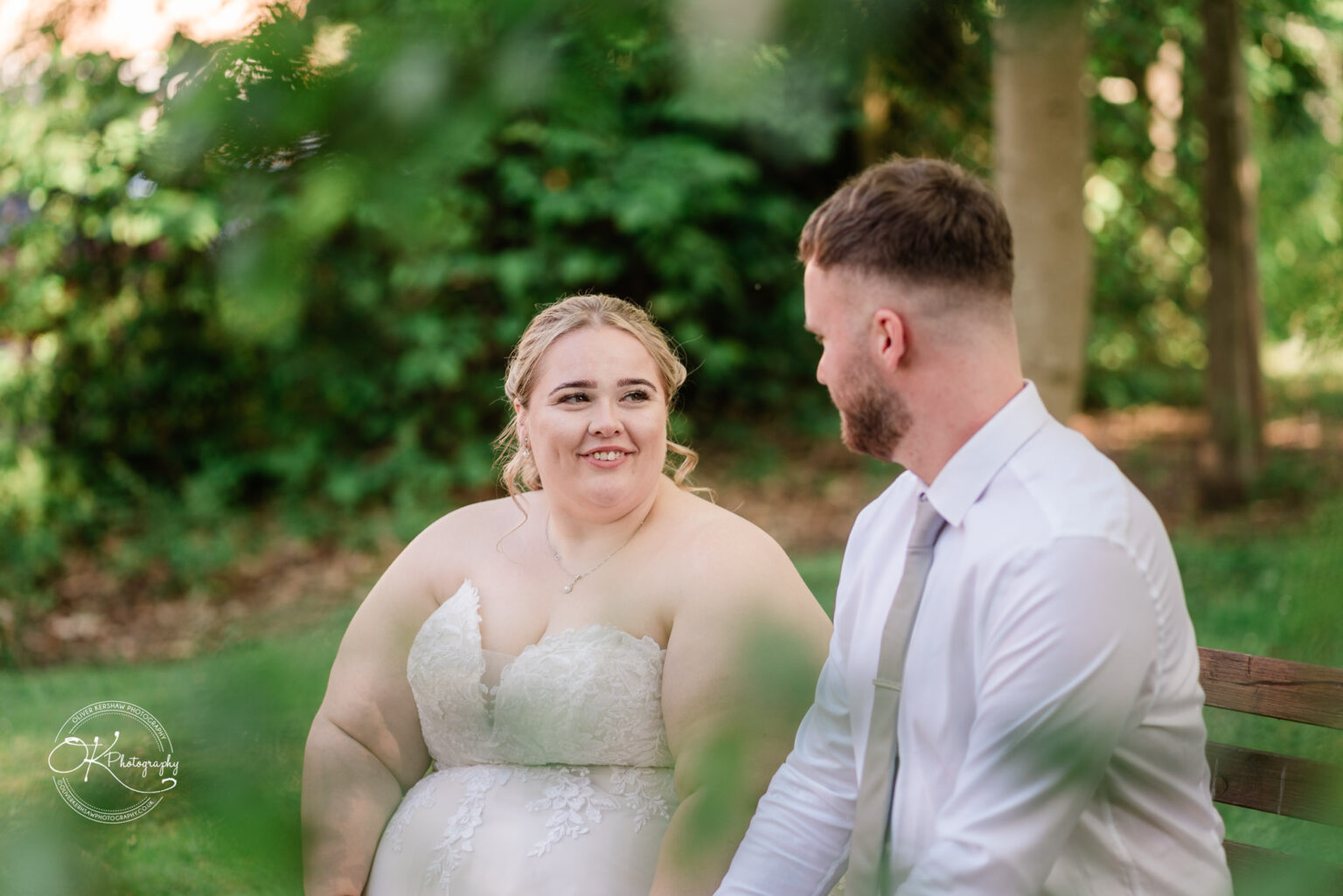 A bride in a lace wedding dress smiles while sitting with a groom in a white shirt and tie, surrounded by greenery.