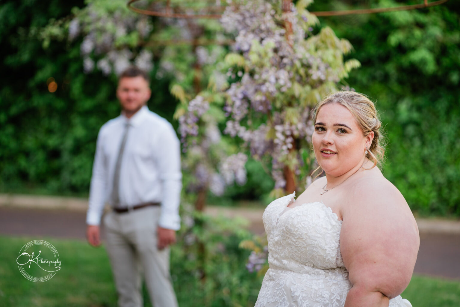 A bride in a white wedding dress poses with a soft smile, while a man in a white shirt stands in the background among flowering greenery.