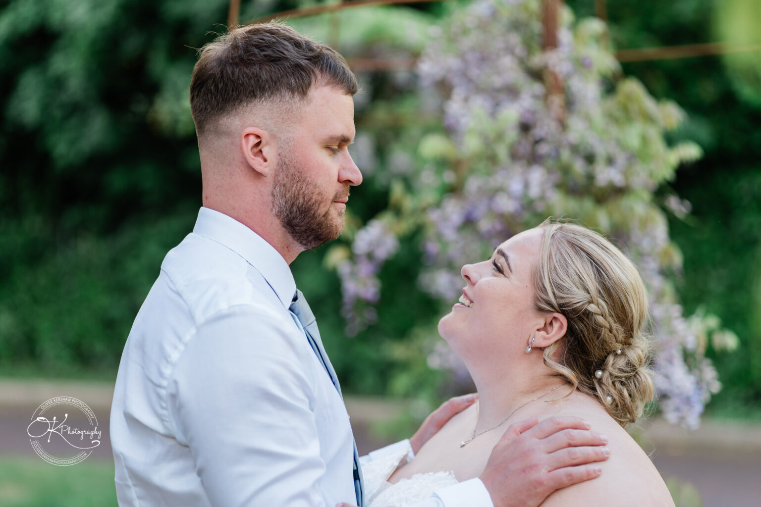 A couple gazes into each other's eyes, with a floral backdrop enhancing their intimate moment. The man wears a white shirt and tie, while the woman, in a strapless dress, smiles warmly.