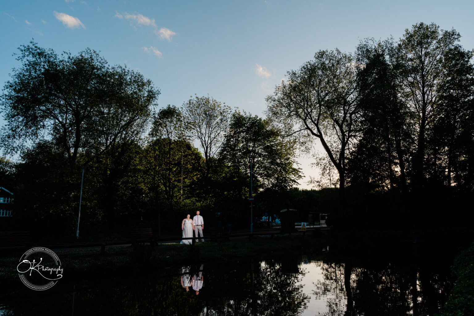 A couple stands by a serene pond at dusk, surrounded by trees reflecting in the water.