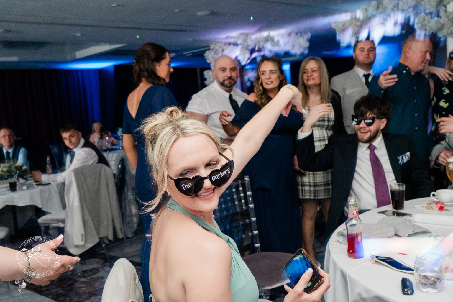A woman in a green dress and sunglasses celebrates at a lively party surrounded by friends and family.
