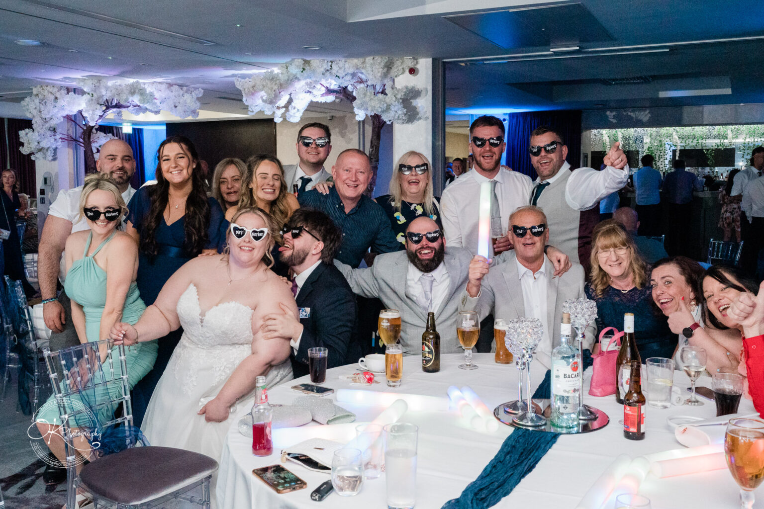 A festive group of people wearing sunglasses, posing cheerfully at a celebration, with drinks on the table and decorative features in the background.
