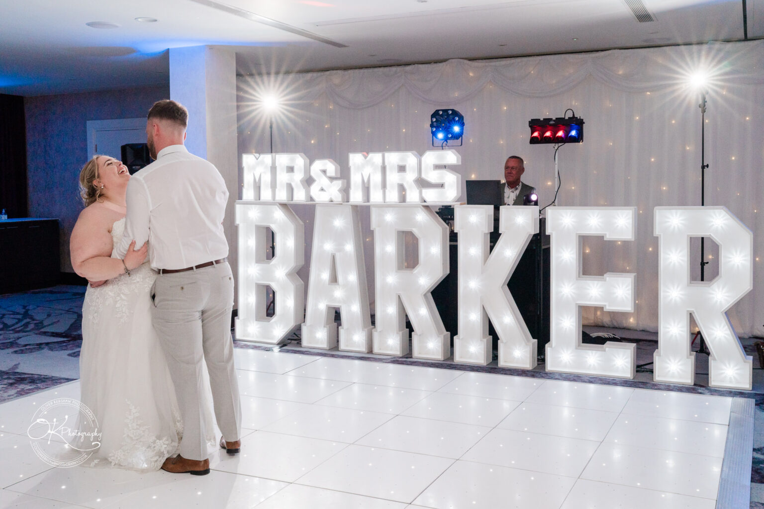 A bride and groom dance together in front of large illuminated letters displaying "MR & MRS BARKER," with a DJ in the background.
