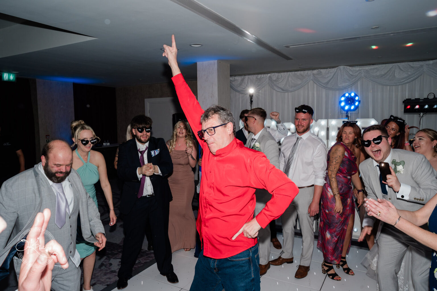 A group of guests dancing at a celebration, with a man in a red shirt striking a playful pose in the centre.