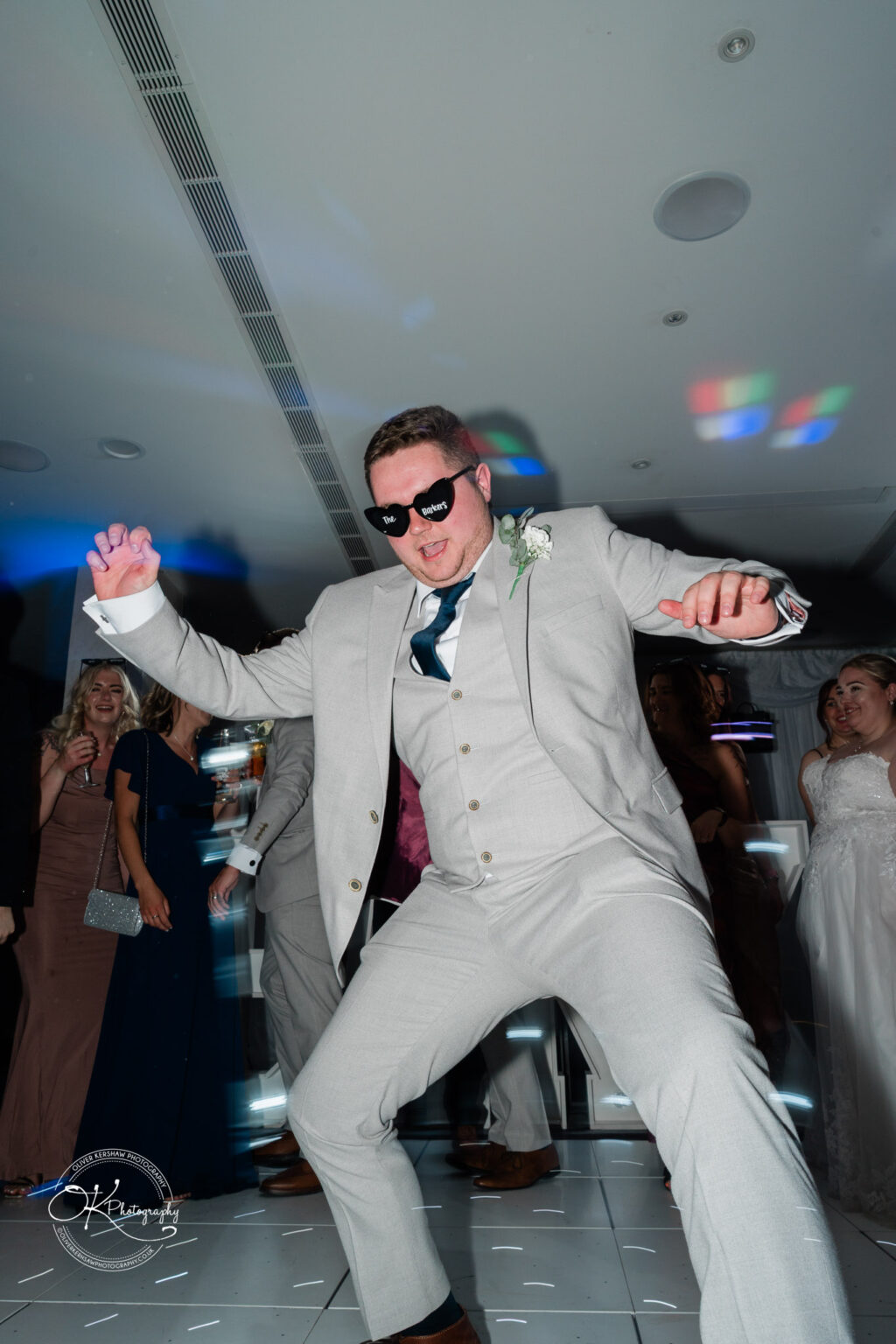 A man in a light grey suit and sunglasses dances energetically on a dance floor, surrounded by guests in formal attire.