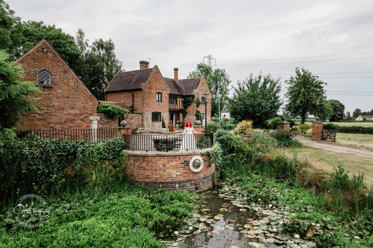 A bride in a white gown stands on a circular stone platform surrounded by greenery and a pond, with a rustic brick farm building in the background.