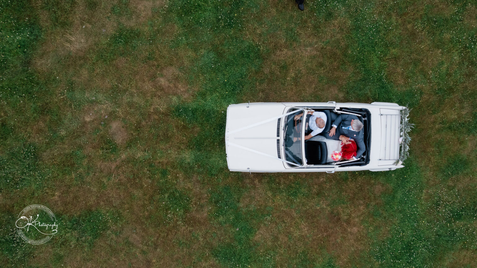 Aerial view of a couple in a white convertible car parked on green grass, surrounded by a serene outdoor setting.