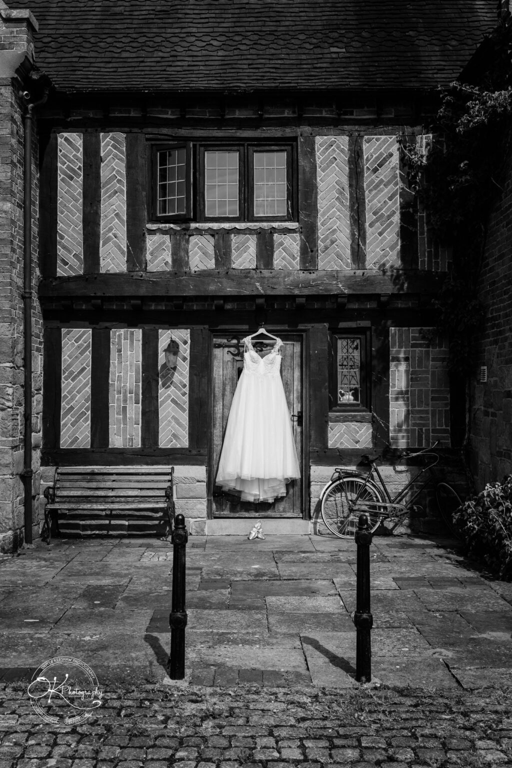 A wedding dress hangs on a wooden door of a rustic brick building, with a bicycle and bench nearby.