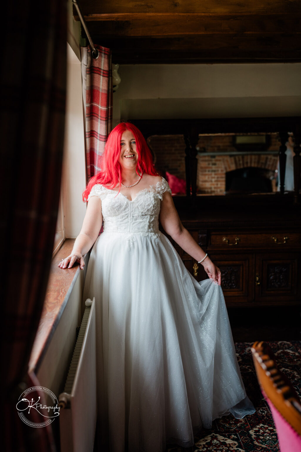 Bride with red hair in a white wedding dress, standing by a window in a rustic setting with wooden accents.