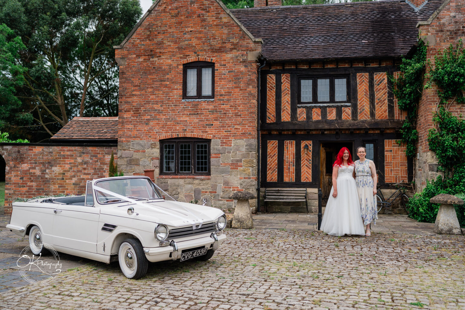 A bride with red hair and her guest stand in front of a rustic brick building, with a white classic convertible parked nearby.