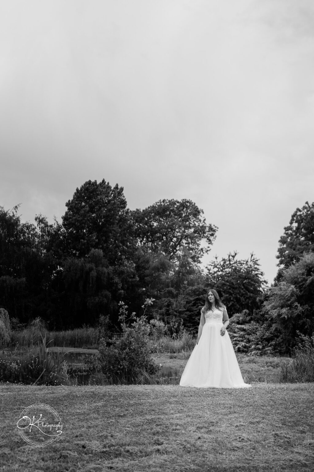 A bride in a flowing white dress walking through a grassy area with trees and a pond in the background, set against a cloudy sky.