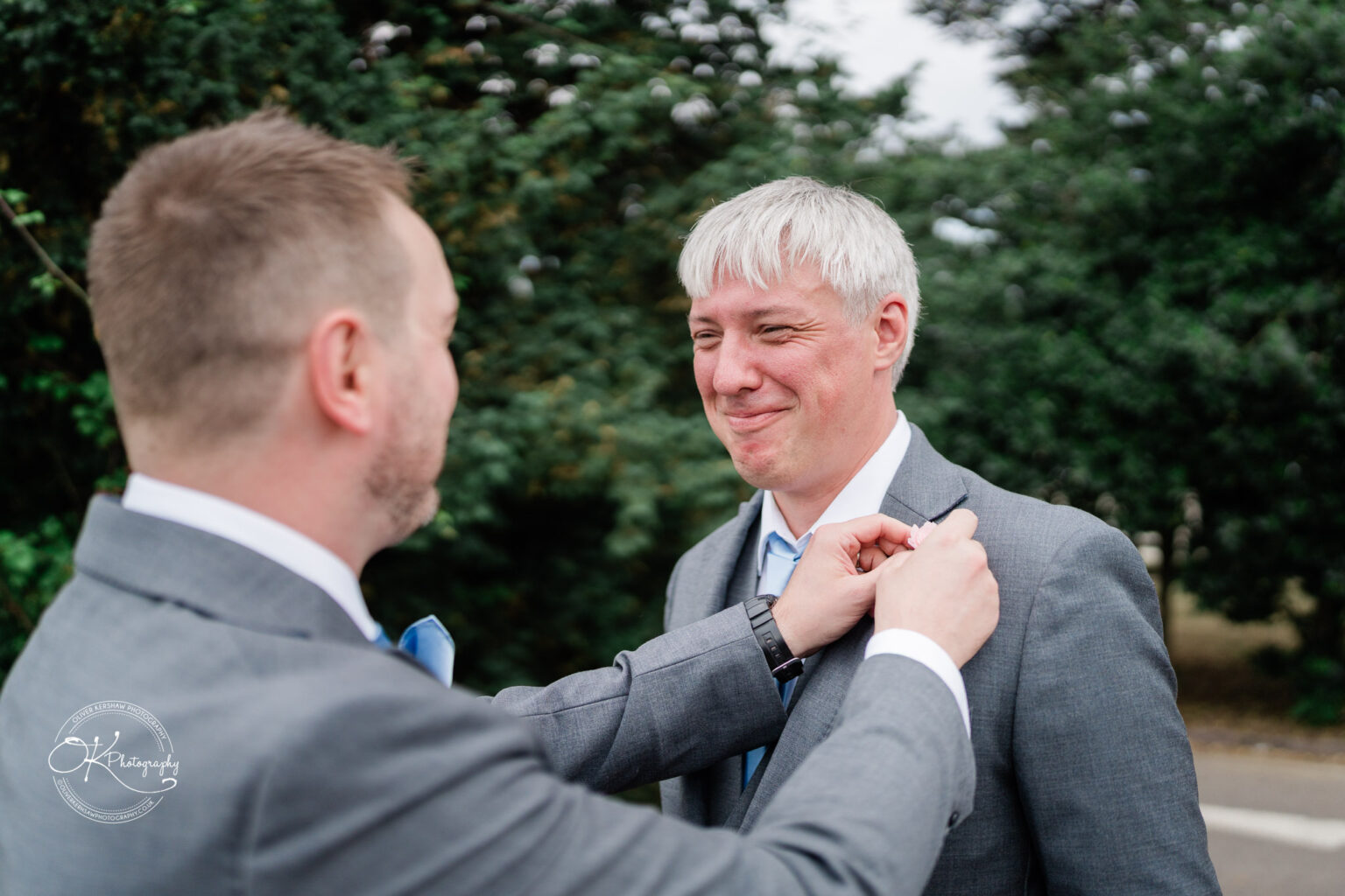 A groom adjusting the boutonniere on the lapel of his best man, both smiling, surrounded by greenery.