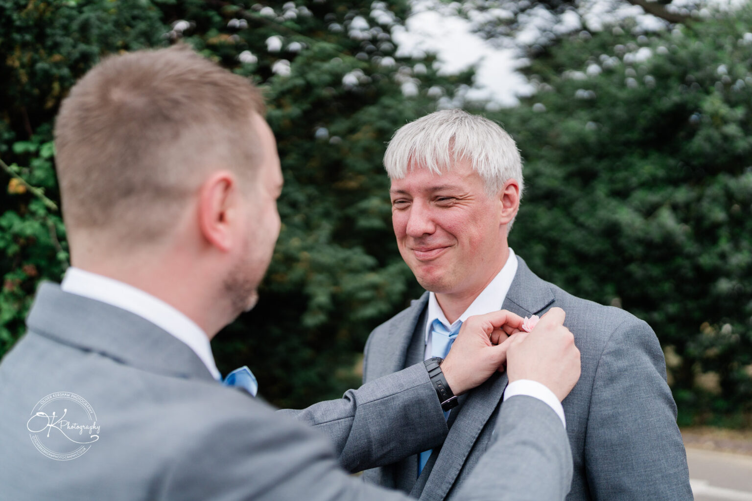 A groom adjusting the boutonniere on a man's lapel, both smiling, surrounded by greenery at Little Markfield Farm.