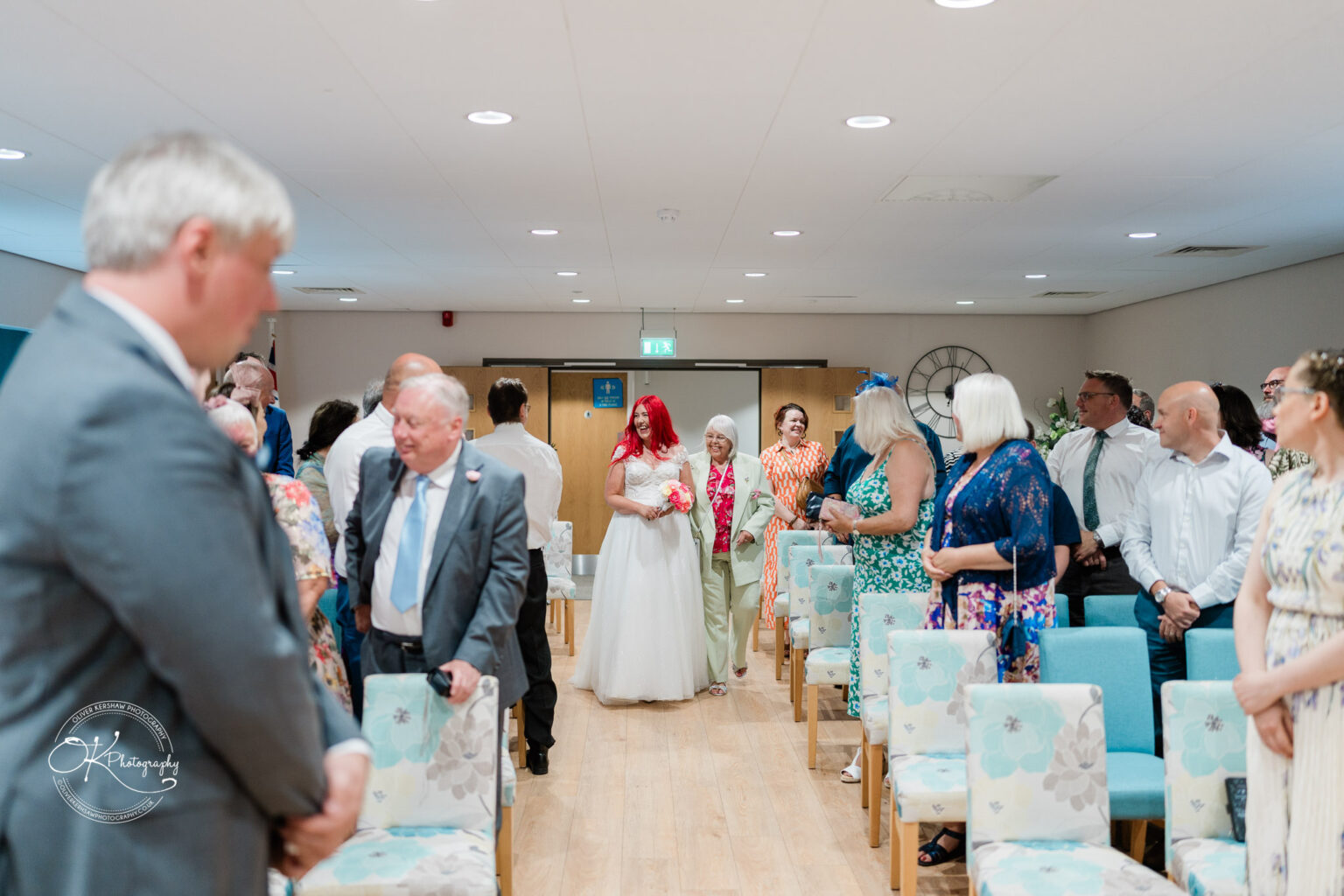 A bride in a white dress and red veil walks down the aisle toward the front, while guests watch from either side in a decorated room.