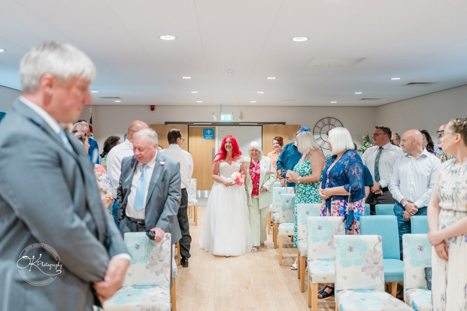 A bride in a white dress and red veil walks down the aisle, flanked by guests seated on either side in a decorated venue.