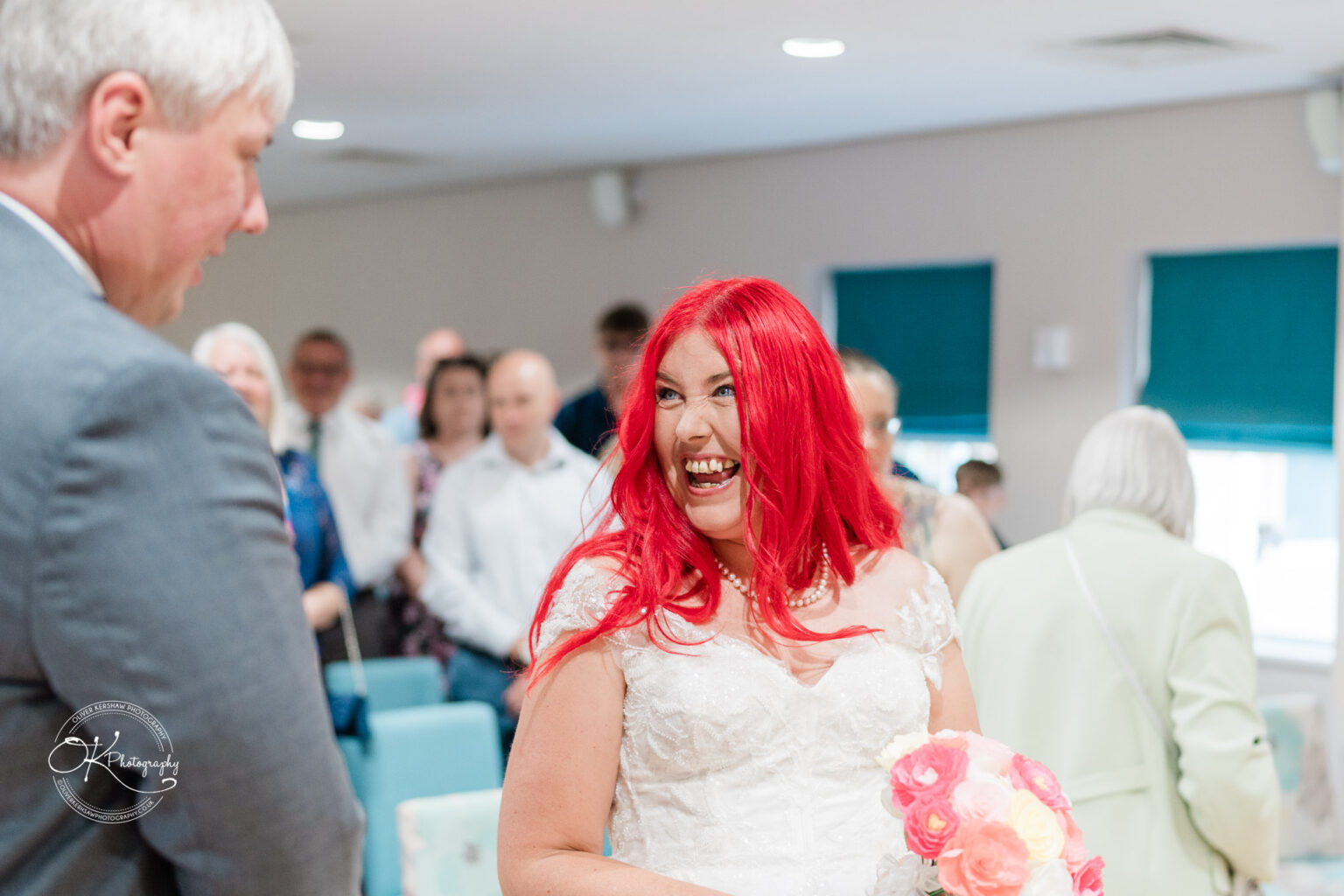 A bride with vibrant red hair smiles joyfully while talking to a guest during her wedding ceremony, surrounded by attendees.