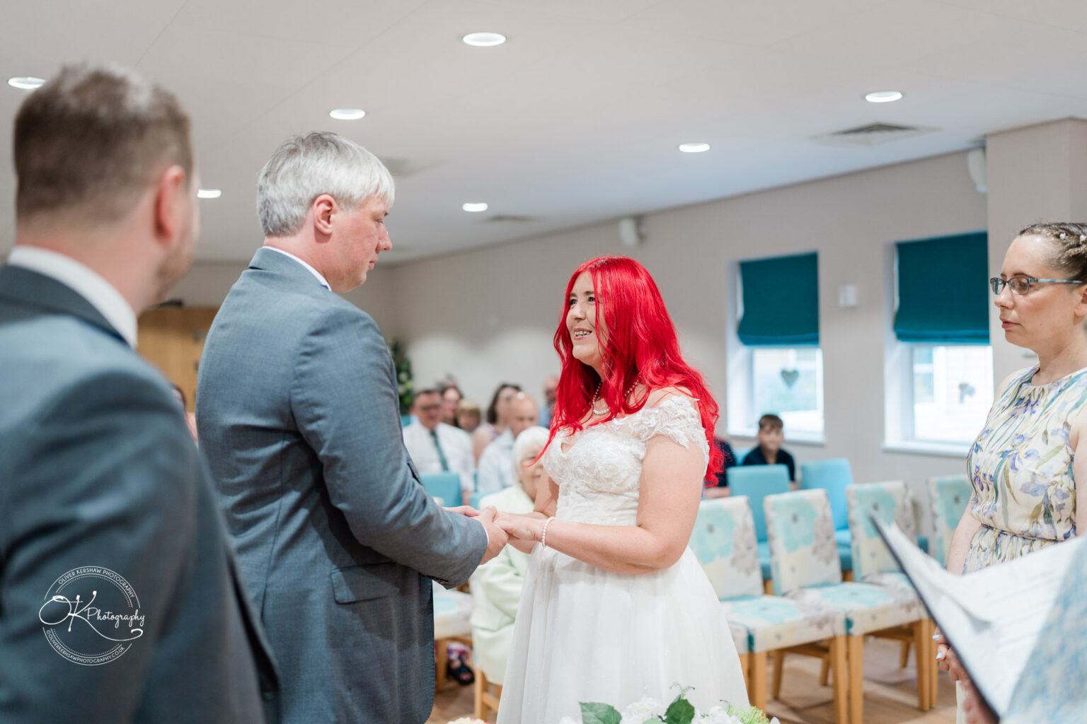 A bride with bright red hair wearing a white dress exchanges vows with a groom in a suit during a wedding ceremony indoors. Guests are seated in the background.