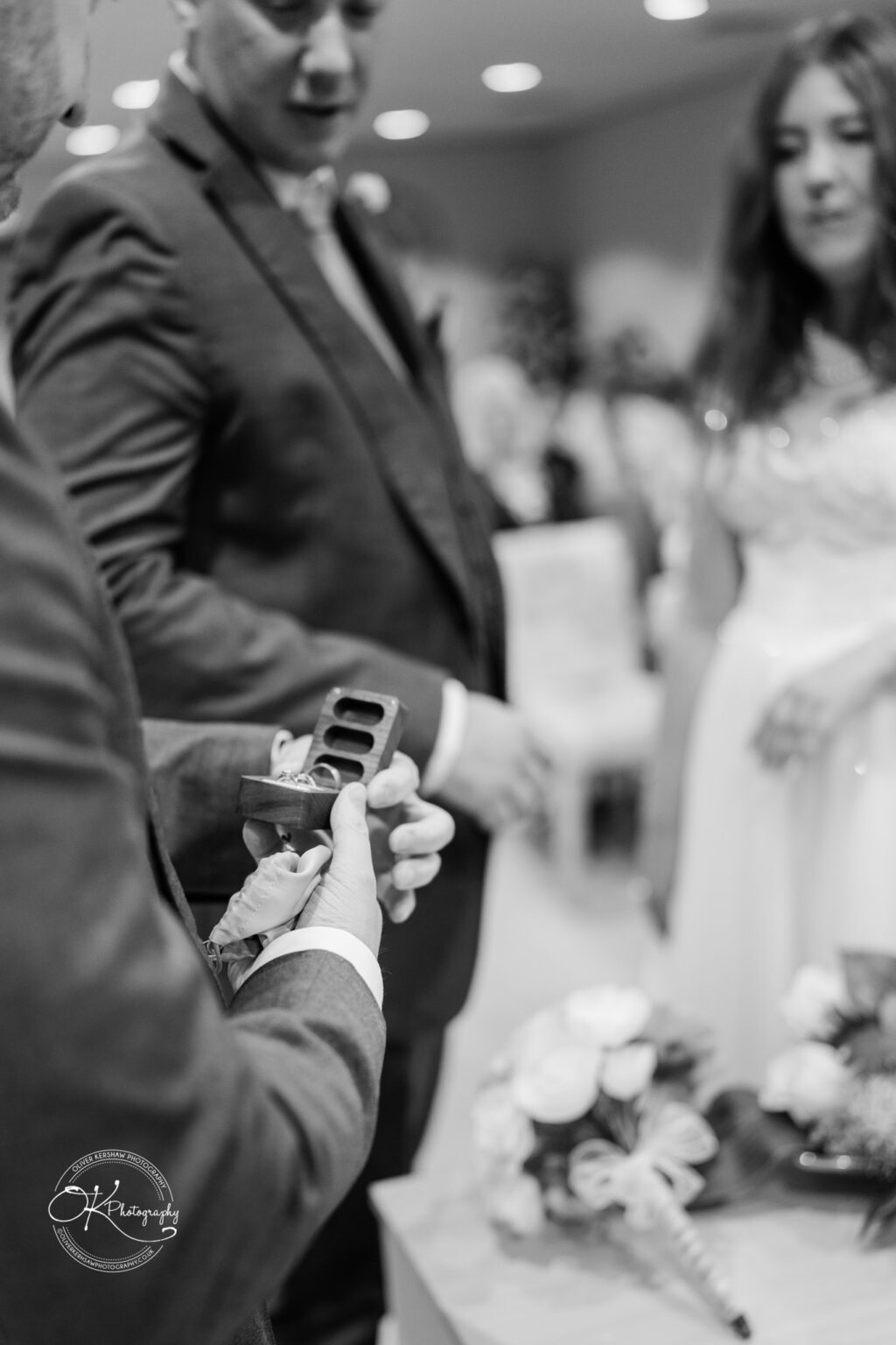 A groom shows a ring in a box to a bride during a wedding ceremony, with guests seated in the background.