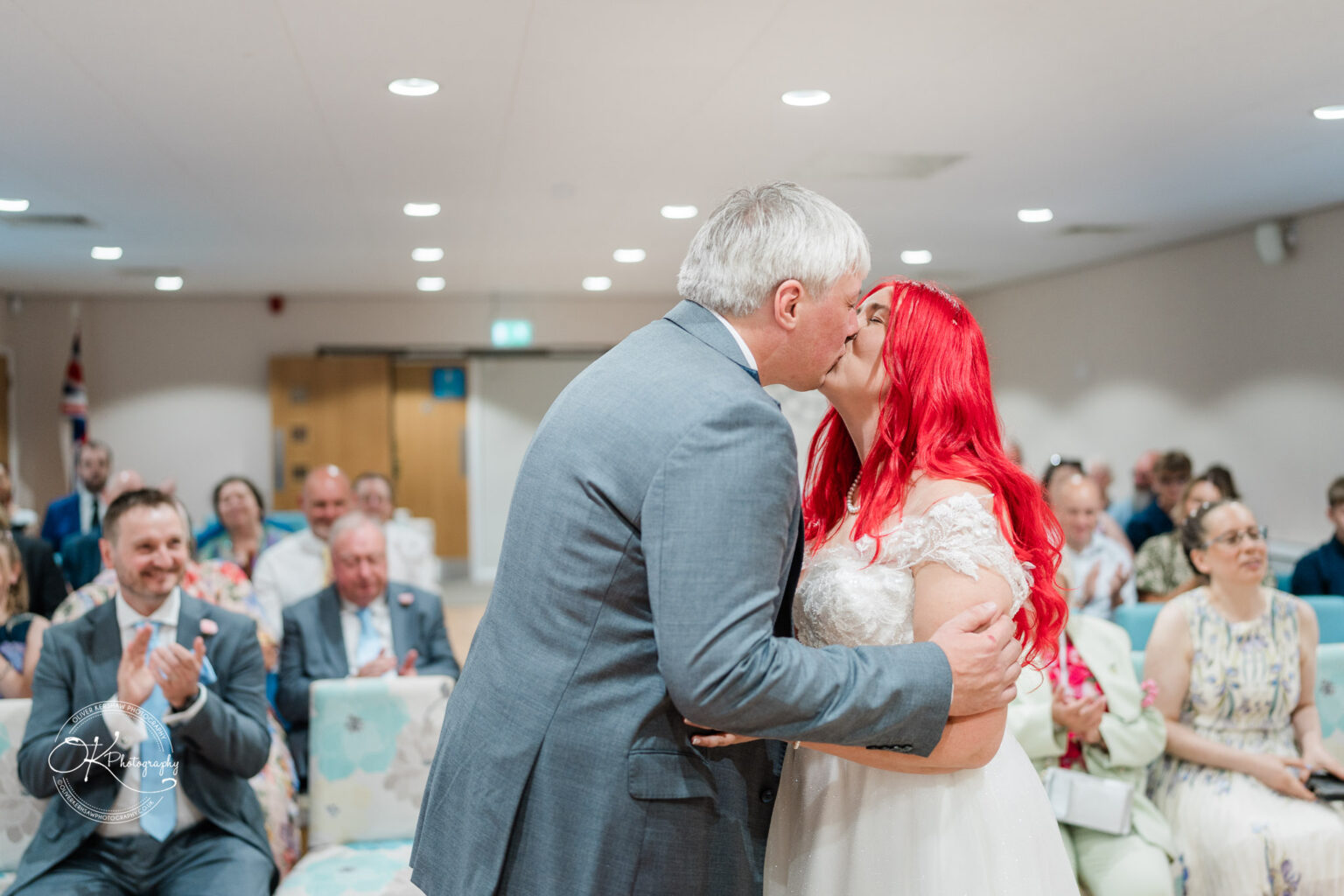 A couple shares a kiss during their wedding ceremony at Little Markfield Farm, with guests applauding in the background.
