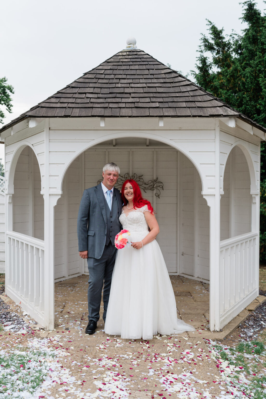 A bride with red hair and a groom, both smiling, stand under a gazebo surrounded by flower petals on the ground.