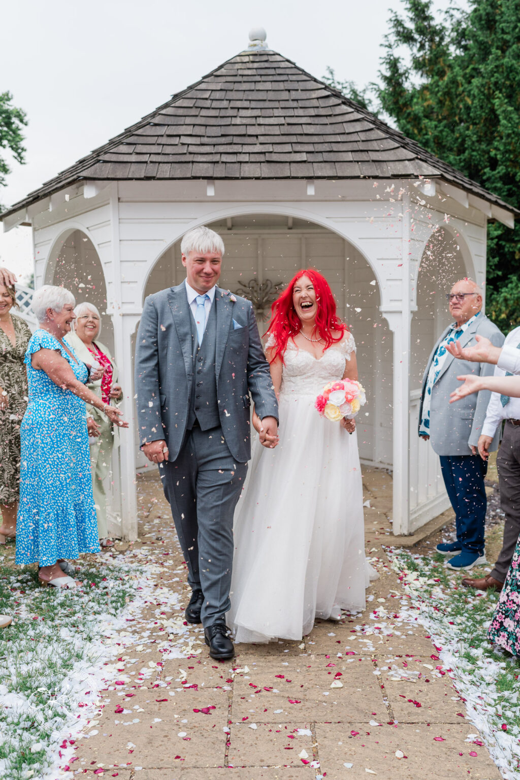 A bride with red hair and a groom in a grey suit walk hand in hand through a flower petal-strewn path, smiling as guests celebrate.