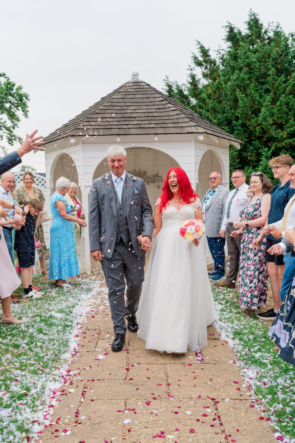 A bride with vibrant red hair and a groom in a grey suit walk hand in hand through a flower petal-strewn path, smiling as guests shower them with petals.