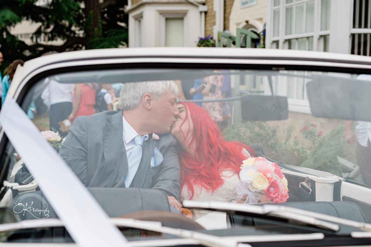 A couple with vibrant hair and a bouquet kiss in a classic convertible, surrounded by wedding guests and greenery.