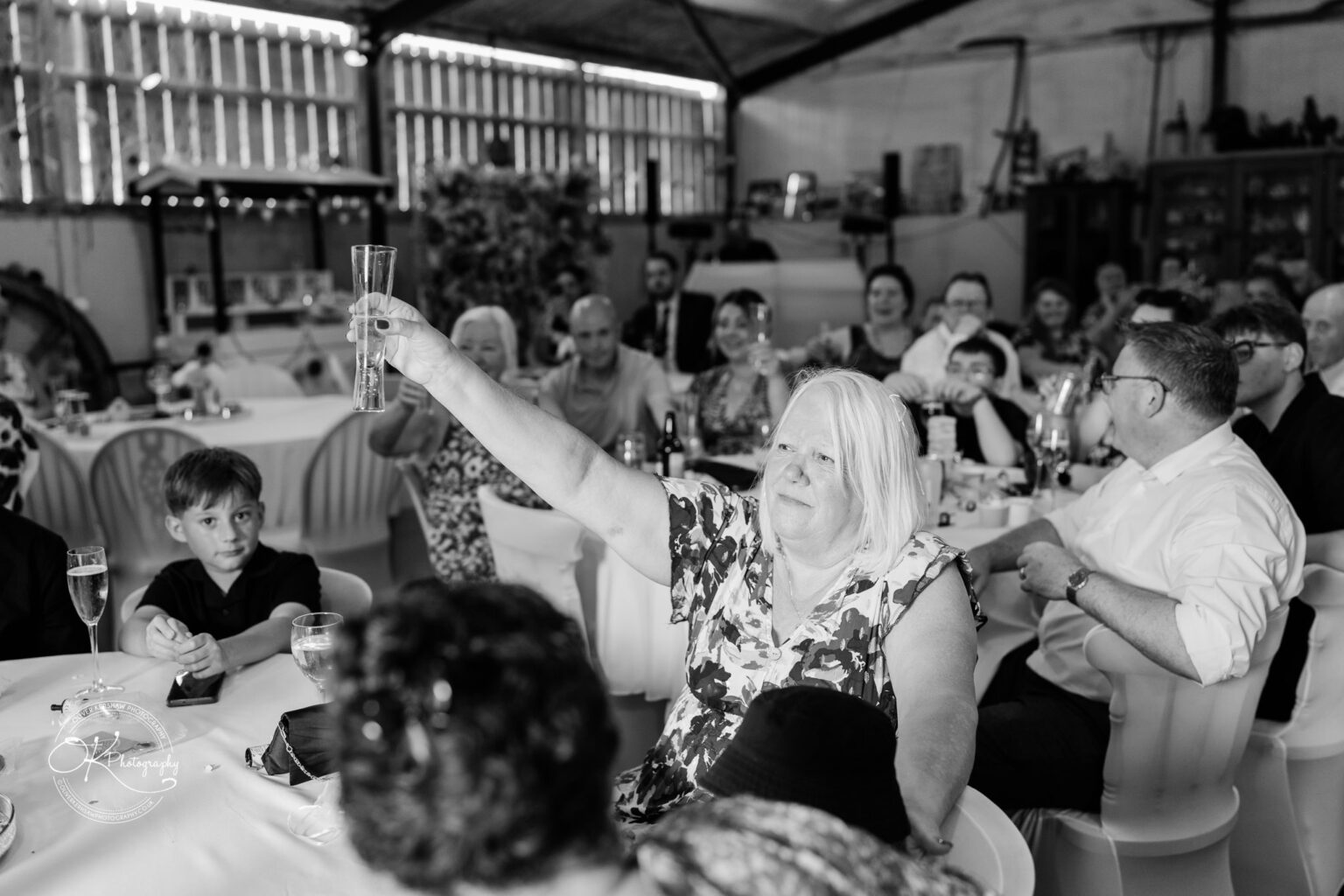 A woman raises a glass for a toast at a wedding reception, surrounded by seated guests in a rustic venue.