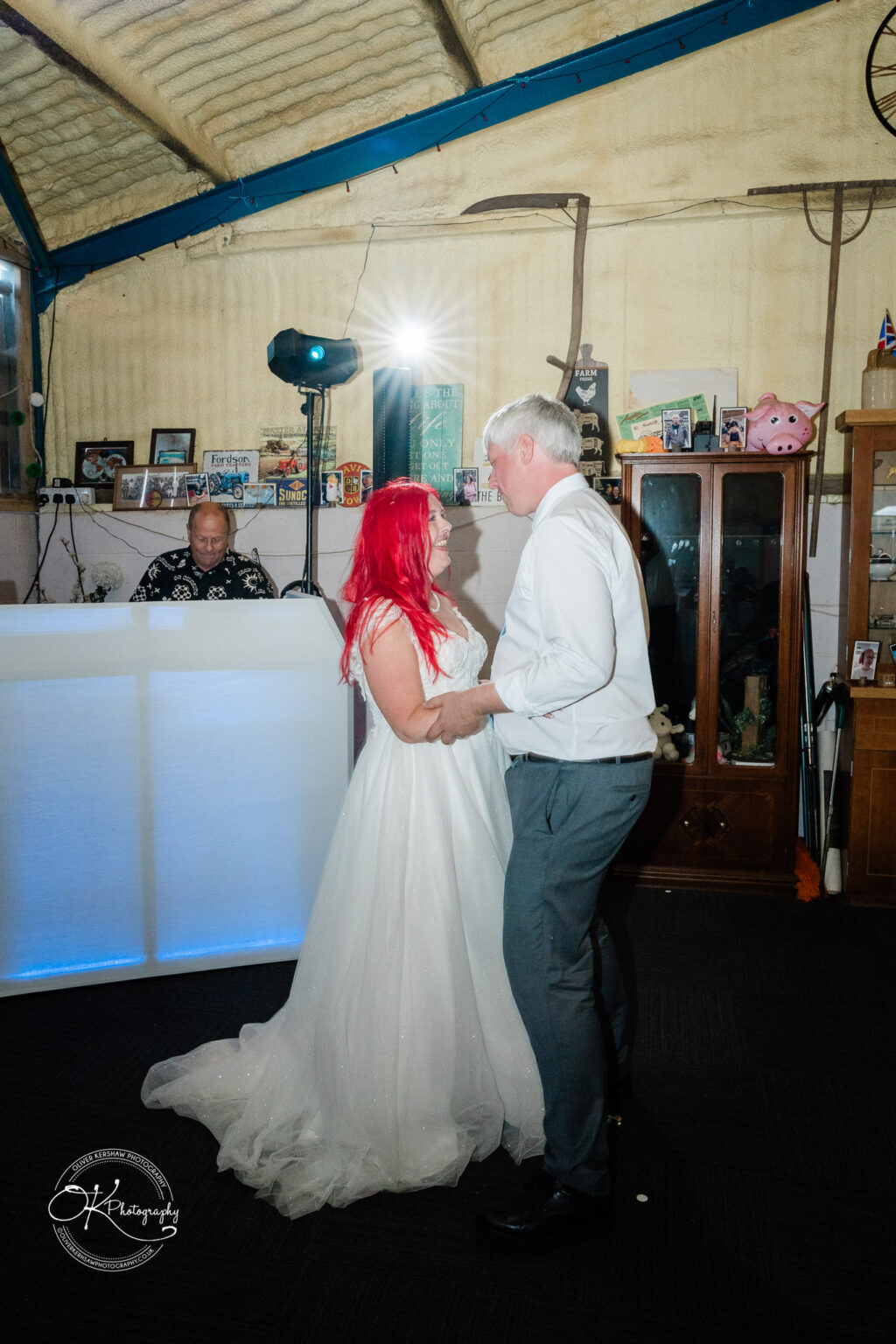 A couple dances together in a rustic setting, with the bride in a white dress and bright red hair, while a DJ operates in the background.
