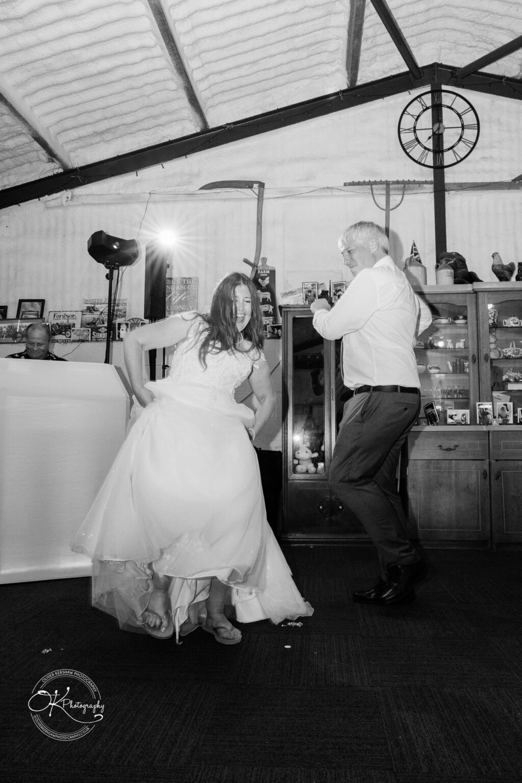 A bride in a white wedding dress dances energetically with a male guest in a rustic indoor venue, filled with decorations.