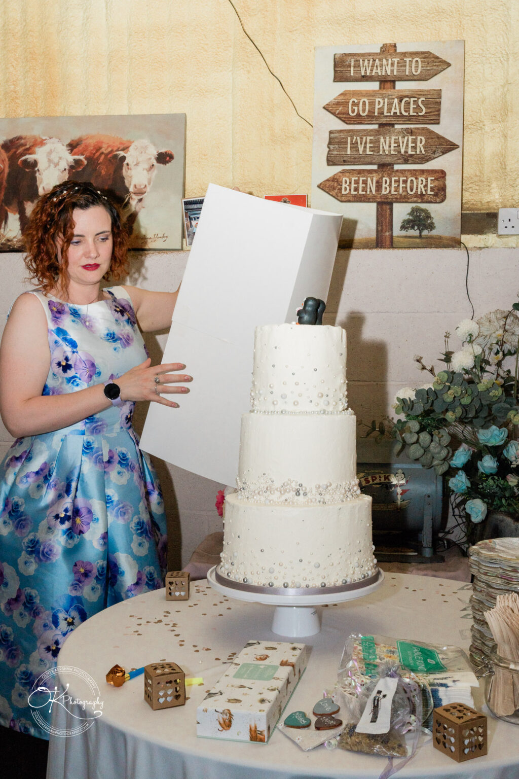 A woman with curly red hair in a floral dress is placing a white box next to a three-tier wedding cake on a table decorated with flowers and party items.