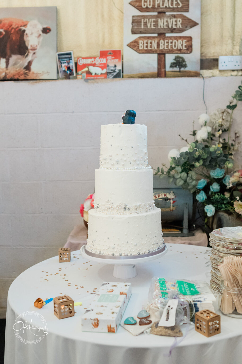 A three-tier wedding cake decorated with white icing, embellishments, and a small figurine on top, set on a table with wedding decor.