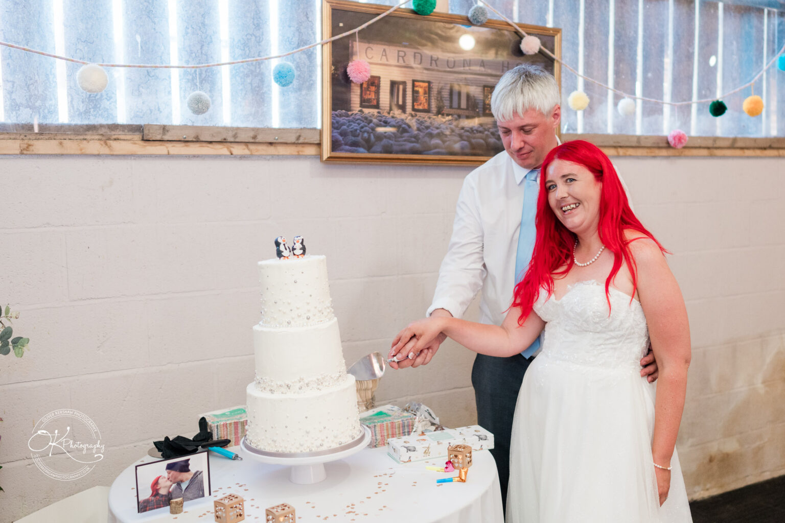 A couple cutting a three-tiered wedding cake, the bride has red hair and wears a white dress, while the groom is in a light blue shirt.