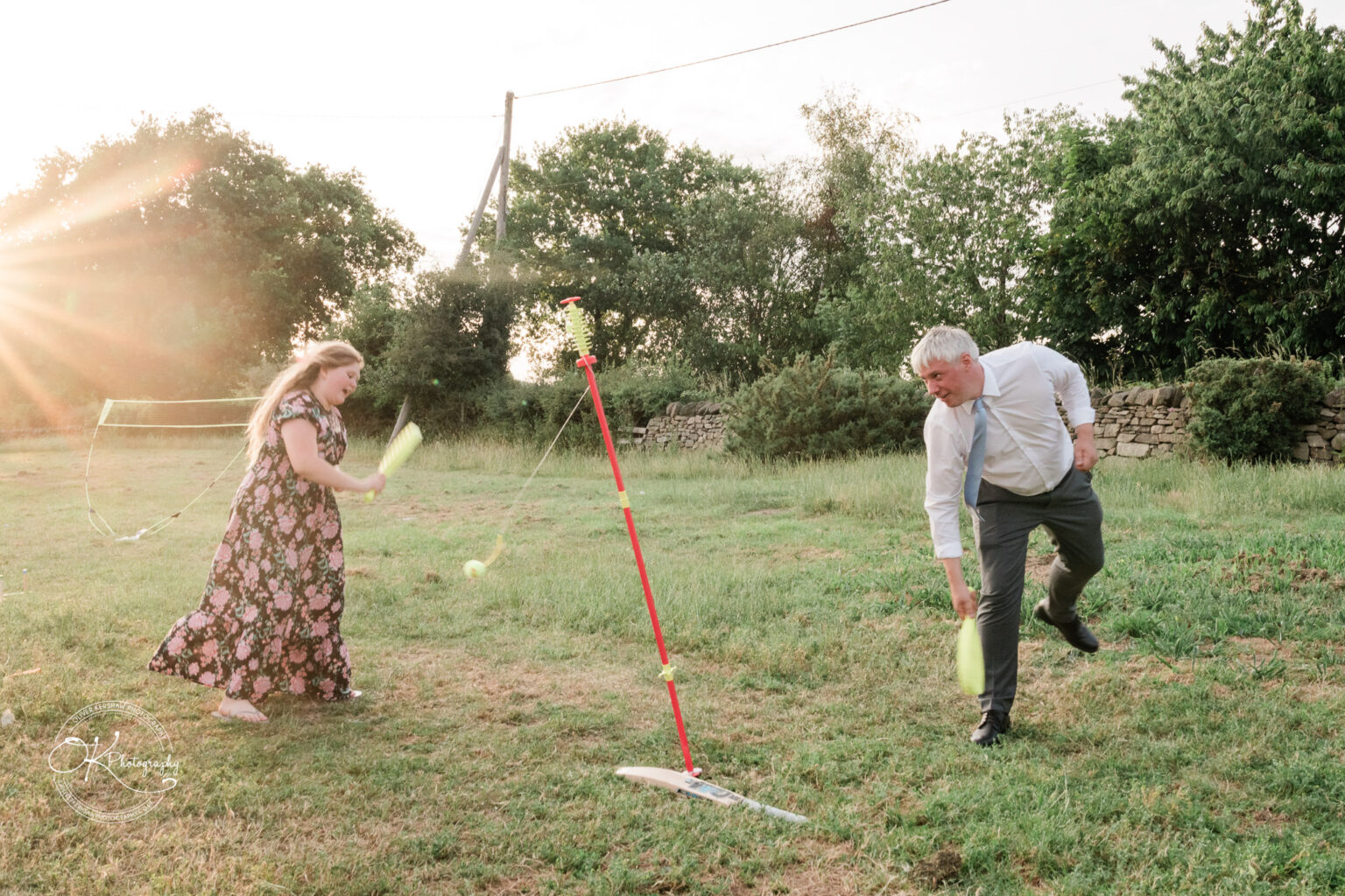 A man in a shirt and trousers plays with a woman in a floral dress, using tennis rackets in a grassy field during sunset.