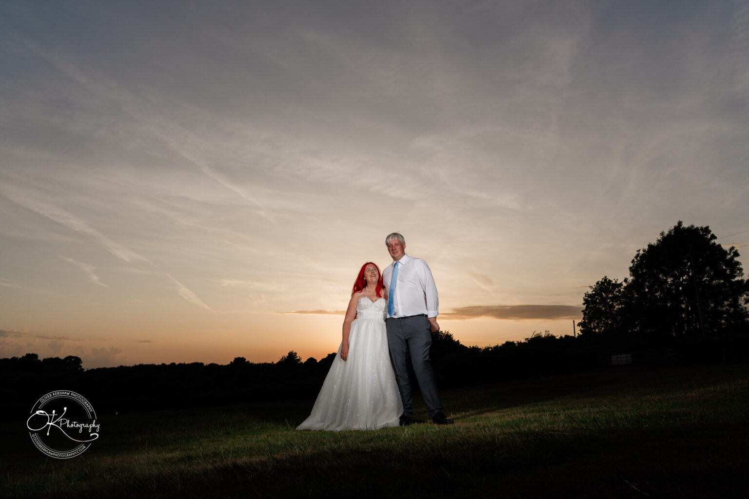 A couple stands together in formal attire against a sunset, with vibrant colours in the sky and a grassy field around them.