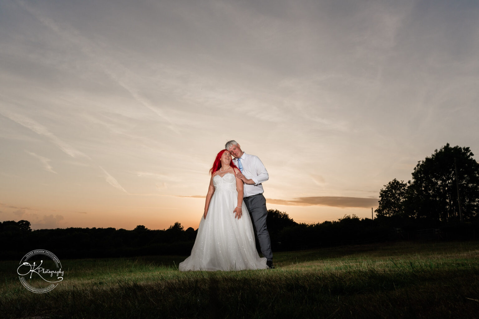 A couple embraces in a field at sunset, with the bride in a flowing white gown and red hair, and the groom in a light-coloured suit.