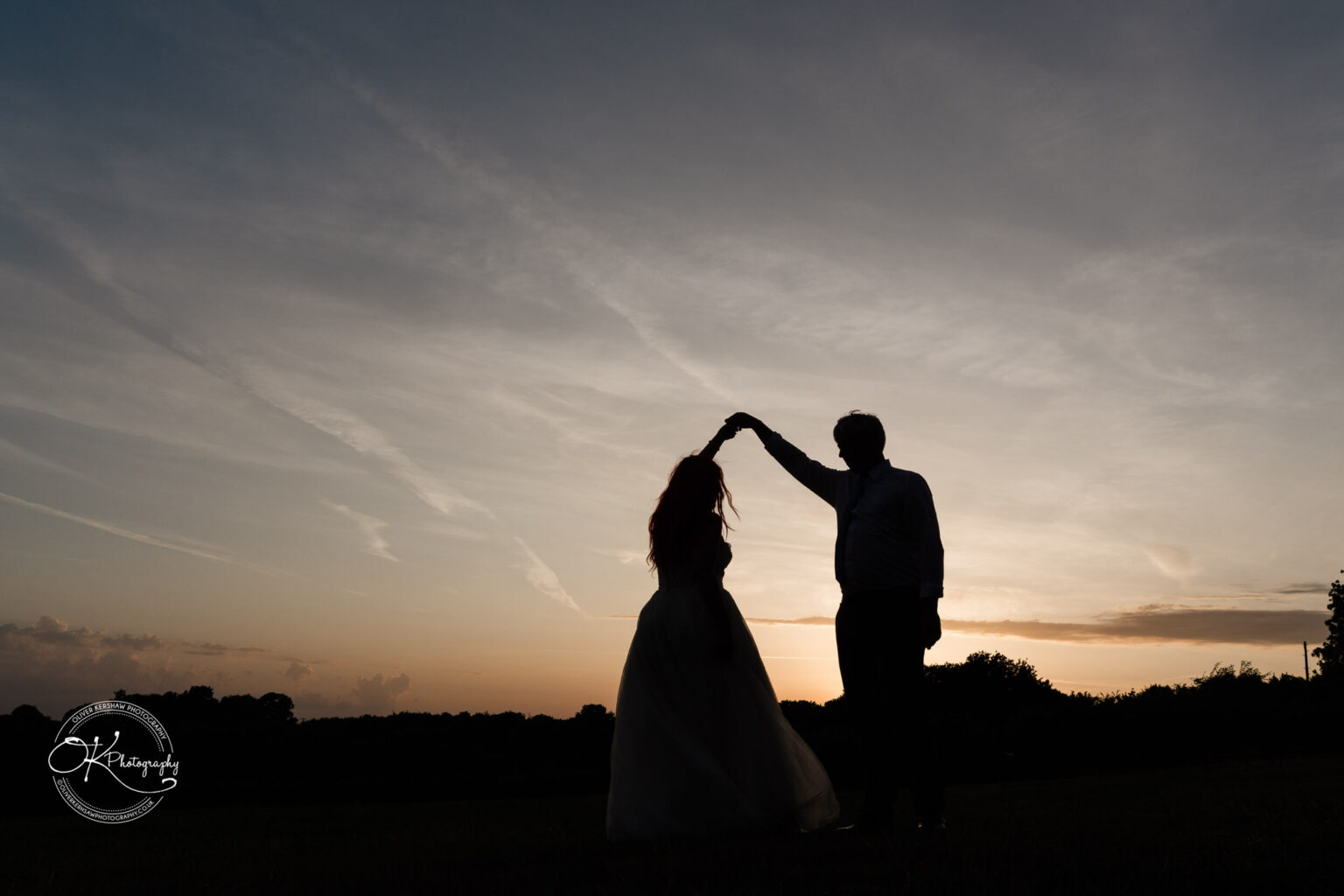 A silhouette of a couple dancing in a field at sunset, with a vibrant sky and trees in the background.