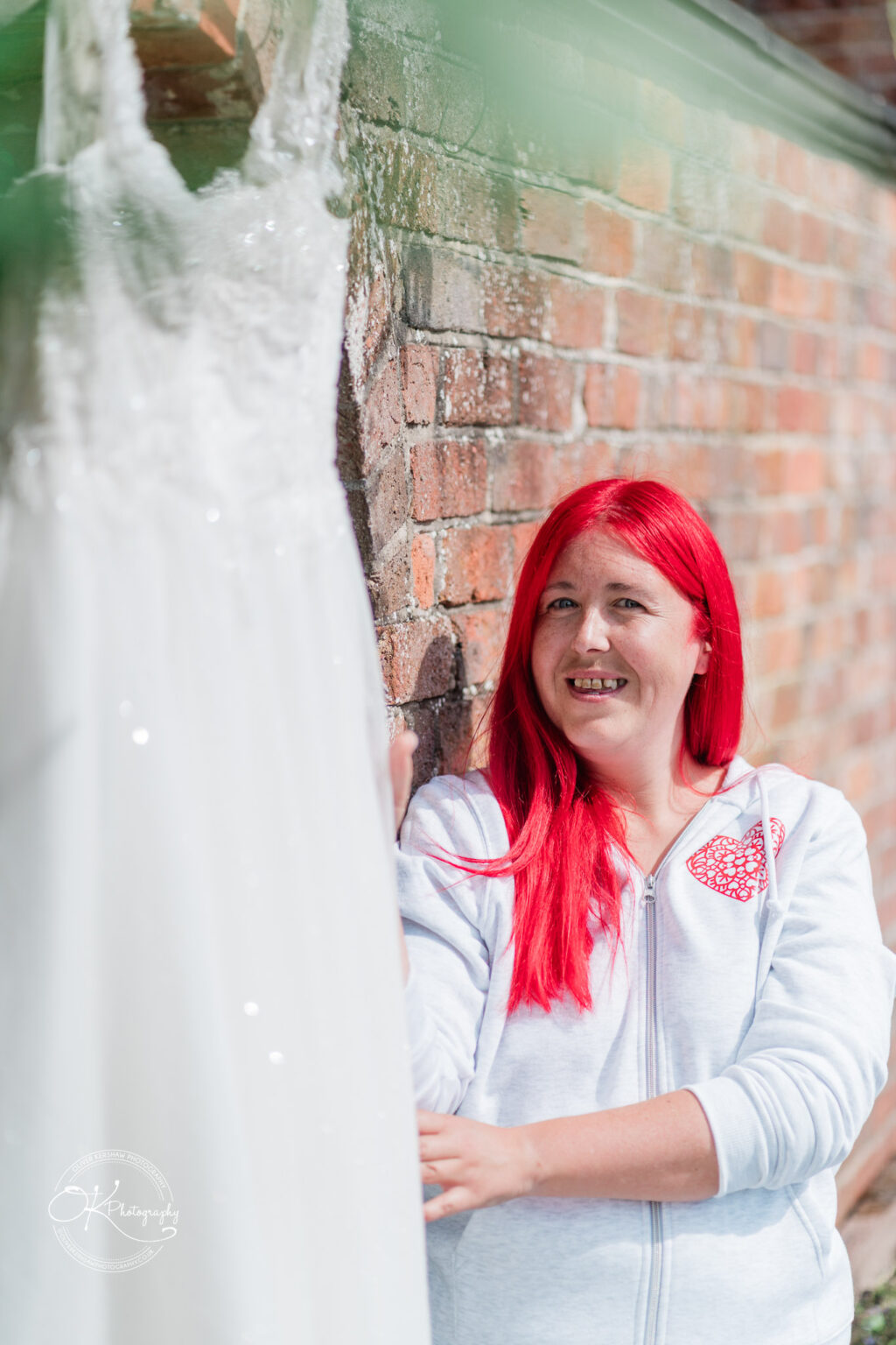 A smiling woman with bright red hair stands beside a wedding dress hanging against a brick wall.