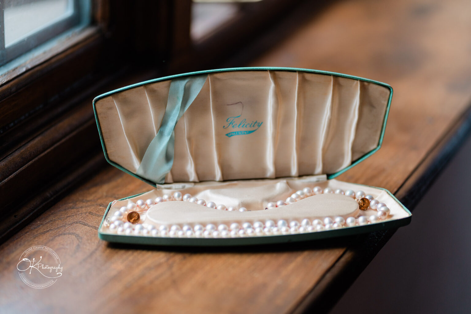 A pearl necklace in an elegant green box, resting on a wooden surface near a window.