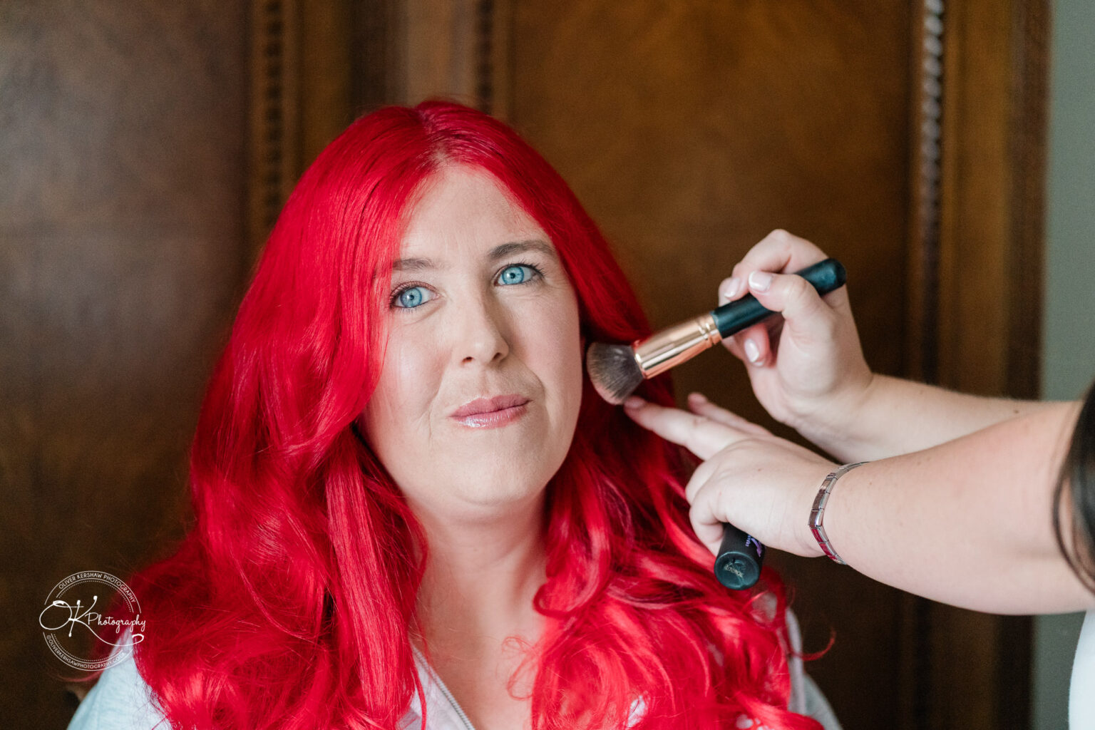 A woman with long red hair is having makeup applied, with a makeup brush near her cheek, and is smiling at the camera.