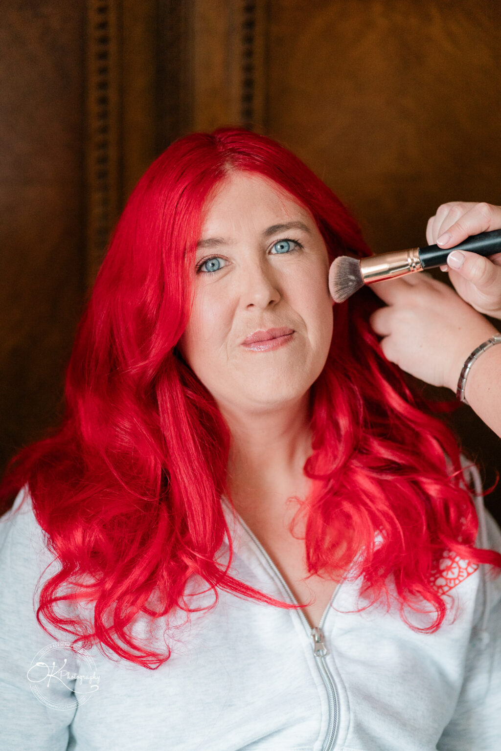 A bride with vibrant red hair is getting her makeup done, sitting comfortably with a stylist applying blush.