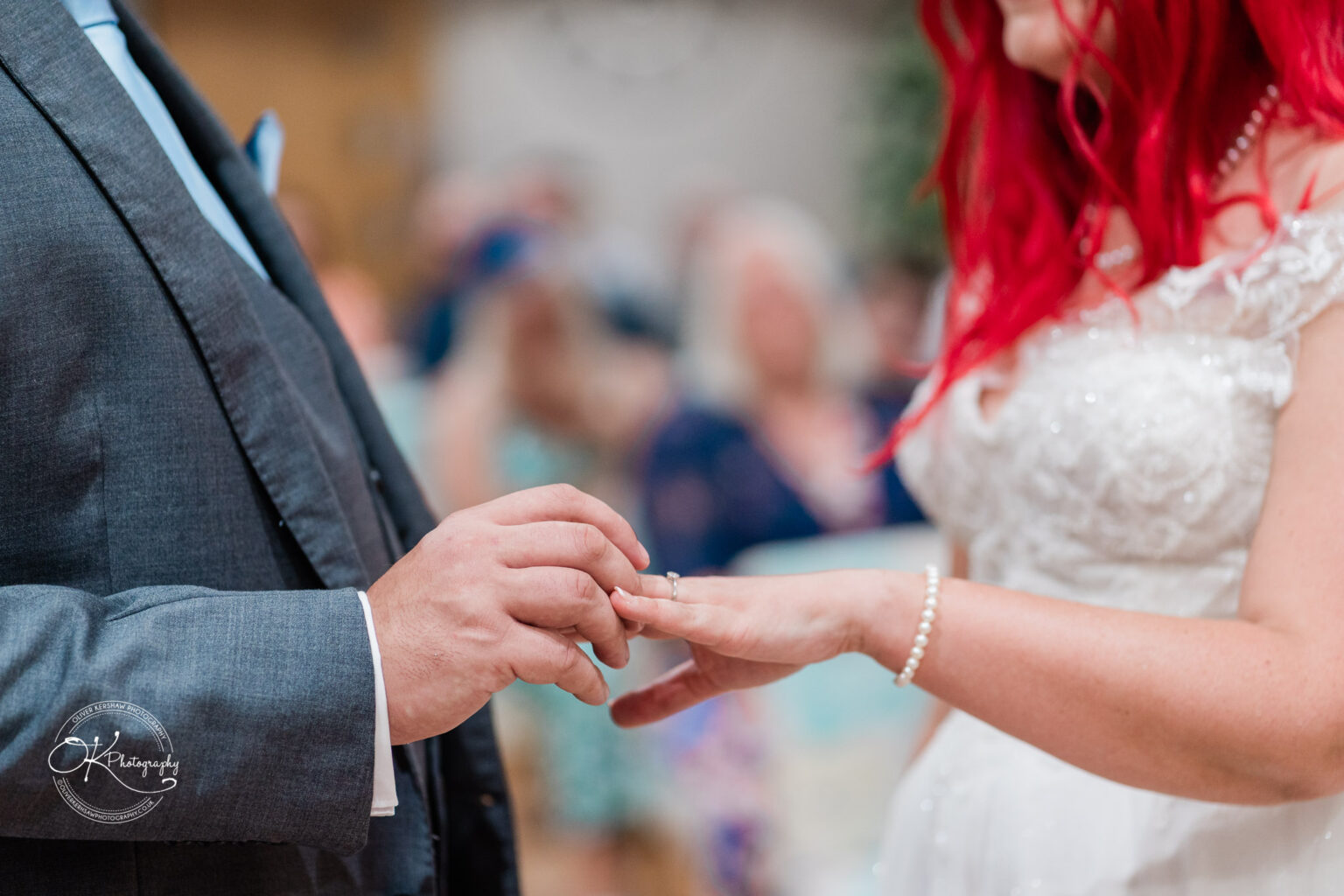 A bride with vibrant red hair and a pearl bracelet delicately places a ring on the groom's finger during their wedding ceremony.