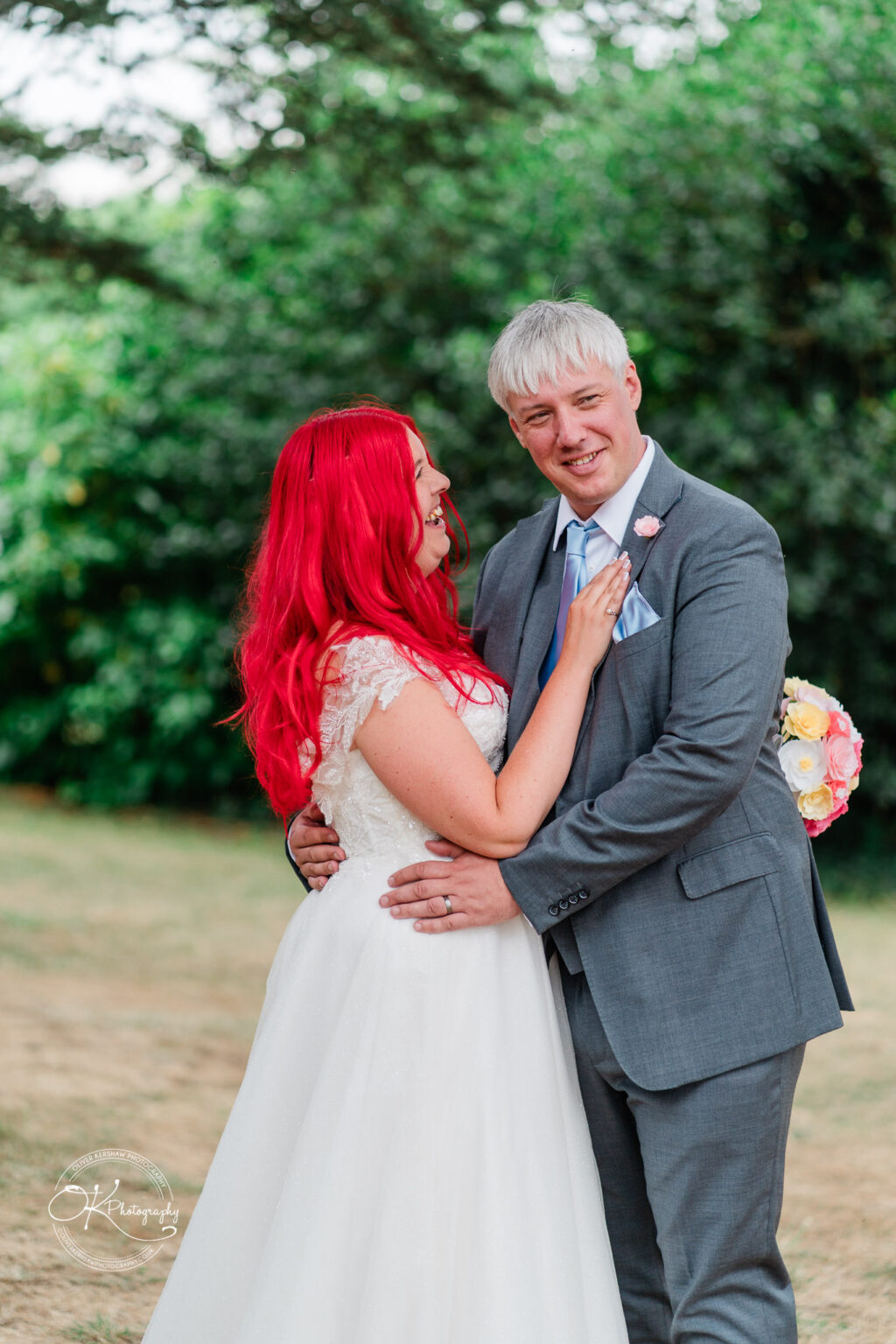 A couple celebrates their wedding, with the bride in a white dress and vibrant red hair, and the groom in a grey suit, smiling happily.