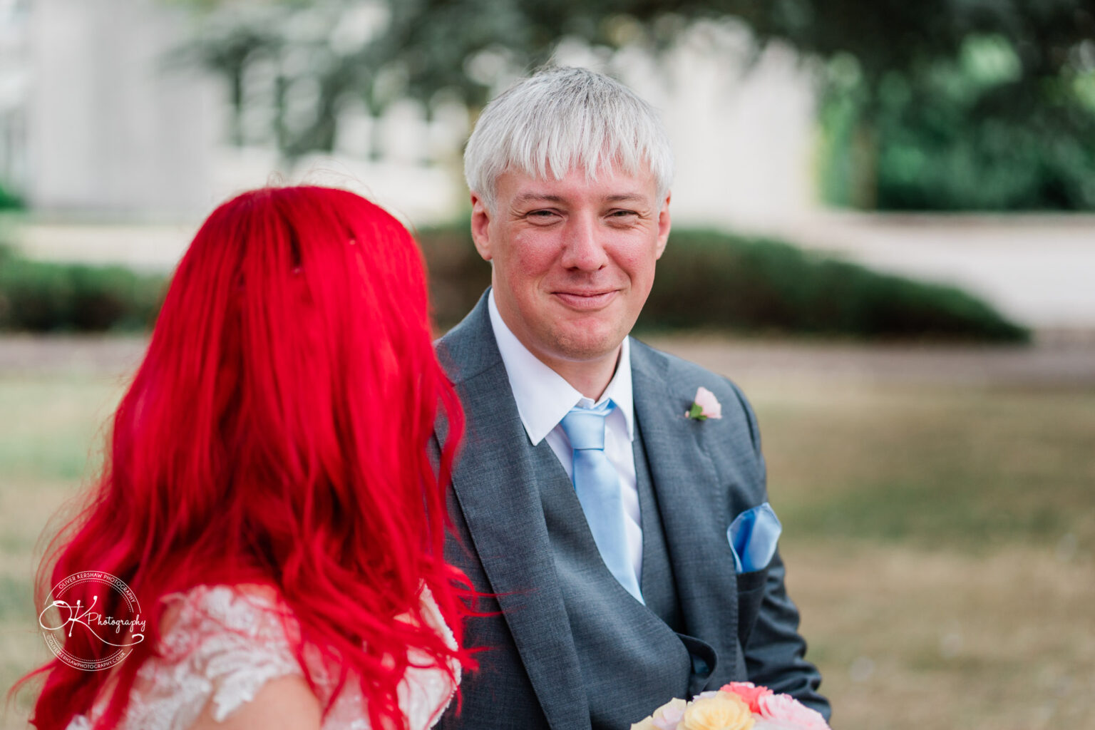 A bride with bright red hair and a groom in a grey suit share a moment outdoors, smiling at each other.