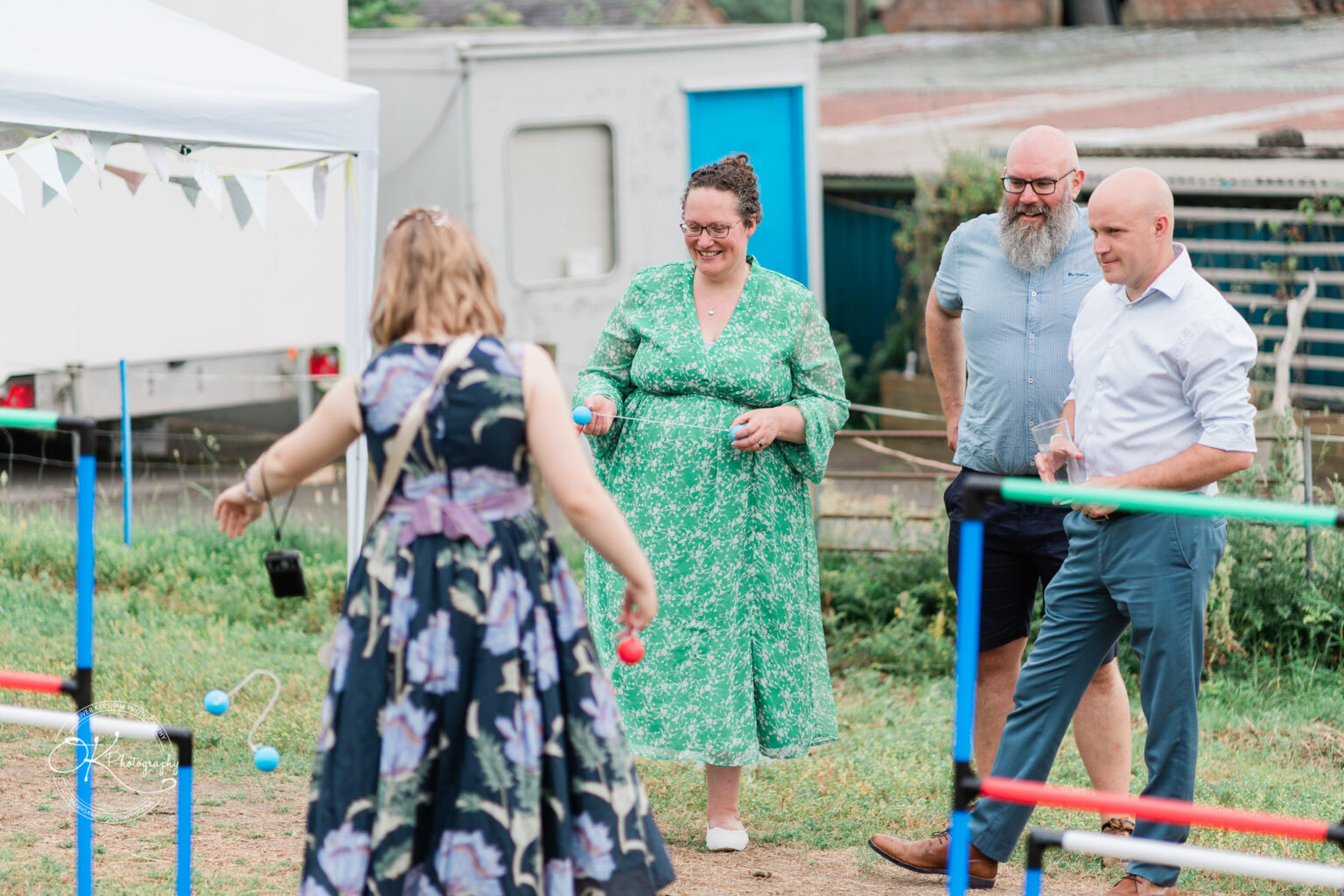 A woman in a floral dress plays a garden game while three attendees observe, smiling, at a wedding reception outdoors.