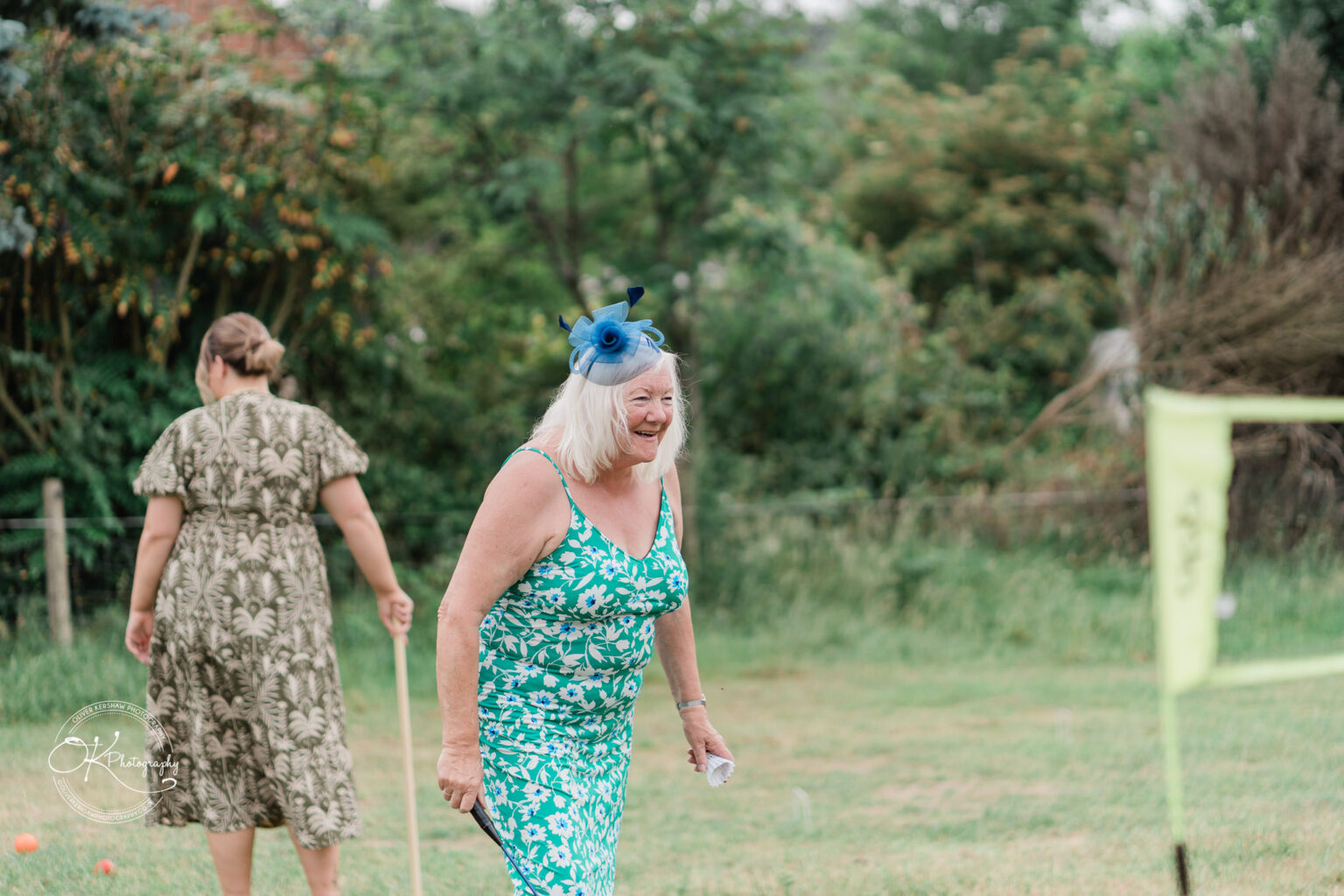 A smiling woman in a green floral dress and blue fascinator walks joyfully across a grassy field, with another person in a patterned outfit in the background.