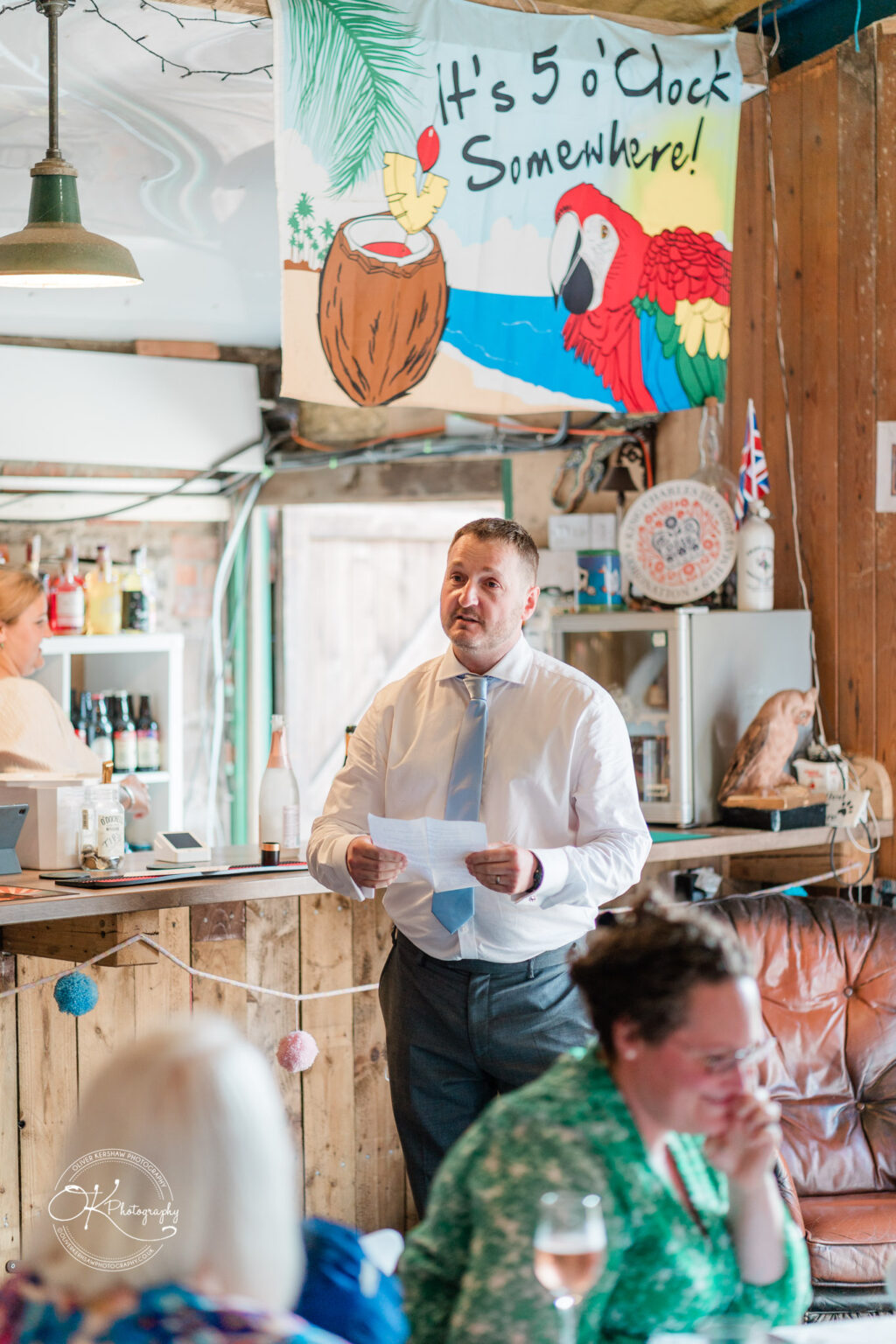 A man in a light blue shirt gives a speech in a rustic bar setting with a tropical-themed sign and a parrot illustration.